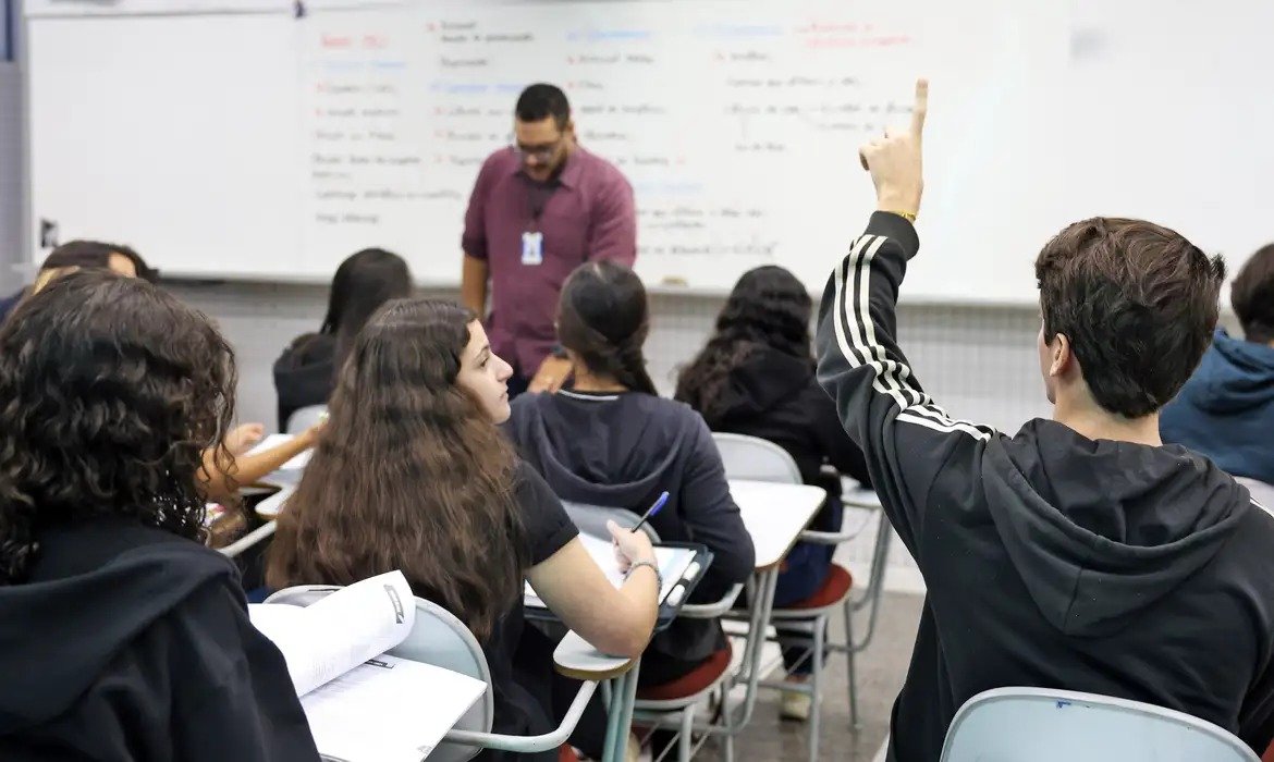 Alunos em uma sala de aula. Professor ao fundo, em segundo plano e aluno, de costas, com a mão levantada.